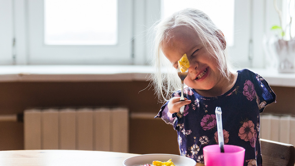 una niña sin brazos y con dos dedos en la mano sostiene con orgullo un tenedor mientras desayuna.