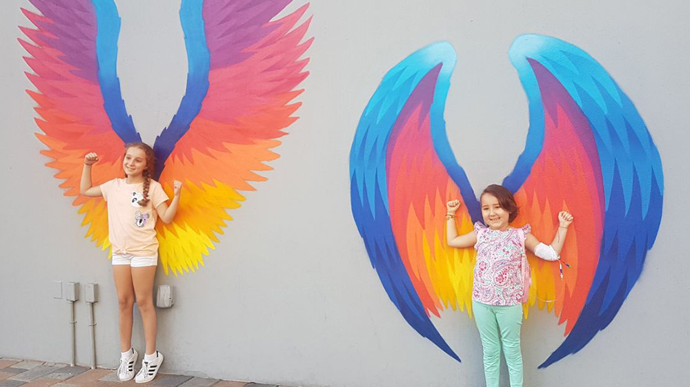 former patient Isabella, and her sister, posing against a wall with multicolor angel wings